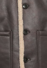 Bruine leren jas met een shearling voering, zwarte knopen en twee zijzakken. De textuur lijkt glad met een zachte binnenkant.