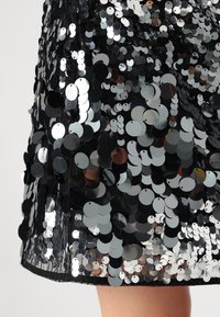 Μαύρη φούστα με παγιέτες που διαθέτει κυκλικές, γυαλιστερές παγιέτες σε διάφορες αποχρώσεις του γκρι, ραμμένες σε μια διάφανη μαύρη ύφασμα. Στολισμένη με σατέν ποδόγυρο.