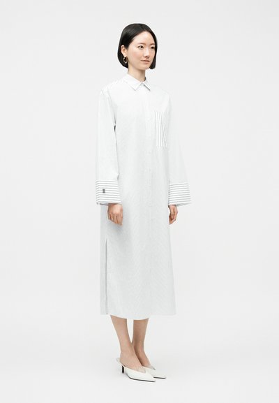 Γυναίκα με κοντά μαύρα μαλλιά, που φοράει μακρύ λευκό πουκαμισοφόρεμα με κάθετες ρίγες, υπερμεγέθη μανίκια και λευκά μυτερά τακούνια, στέκεται μπροστά σε λευκό φόντο.