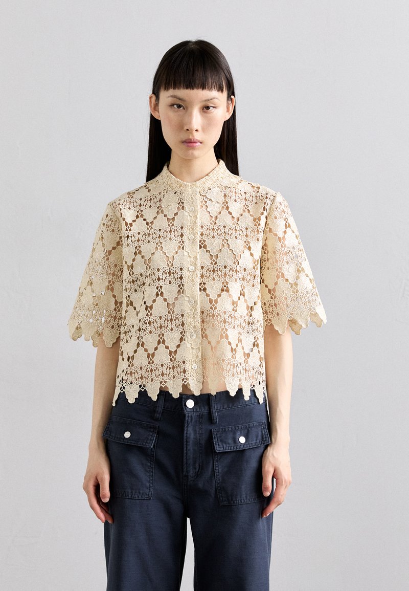 Šviesiai beige spalvos, trumpomis rankovėmis, nėriniuota bluzonė su banguotais kraštais ir mygtukų užsegimu priekyje. Sukurta su gėlių raštu ir permatomu audiniu.