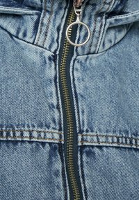 Džínová bunda se zipovým zapínáním, světle modrá barva, vybledlá textura, kontrastní prošívání a kruhová táhla na zipu.