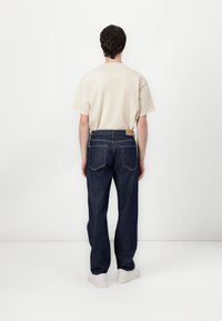 Jeans en denim bleu foncé avec une coupe droite, cinq poches et une étiquette en cuir tan à l'arrière, associés à un t-shirt beige et des baskets blanches.