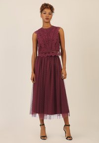 Kleid aus Spitze und Tüll in tiefem Burgunderrot. Es hat ein eng anliegendes Spitzen-Oberteil mit gewellten Kanten und einen fließenden Tüllrock mit Punkten.