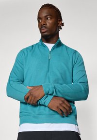 Teal värvi neljandik-zip pullover tekstuuriga kangast, millel on kõrge krae ja pikad varrukad. Valge särk on all, ribatud varrukaotsad ja äär.