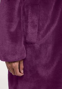Abrigo de pelo morado y suave que presenta una textura blanda, bolsillo visible y un ajuste relajado con mangas ligeramente ajustadas.