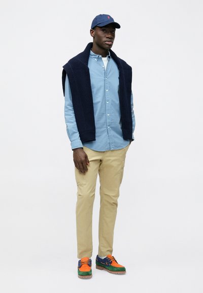 Camisa azul clara de botão, suéter de malha azul-marinho colocado sobre os ombros, calças bege e ténis multicoloridos. Boné simples completa o visual.