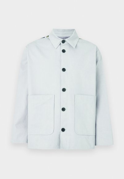 Veste en coton bleu clair avec col pointu, boutons noirs, deux poches avant et légers accents aux épaules. Design simple et fonctionnel.