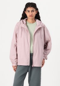Lys pink, rutete vindjakke med hette, elastiske mansjetter og glidelås foran. Bært over en lys grønn skjorte og grå bukser.