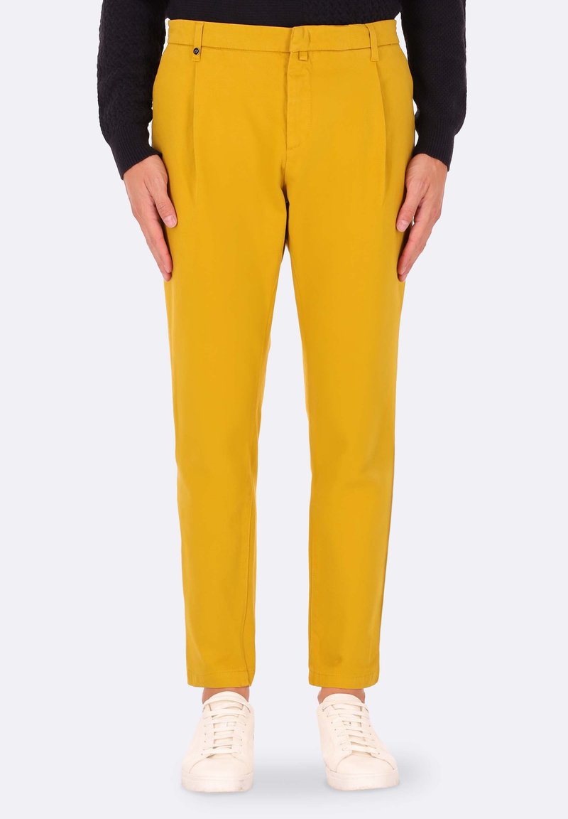 Pantaloni gialli con una vestibilità sartoriale, frontale piatto e tasche laterali, abbinati a sneaker bianche. Il materiale sembra liscio e leggero.