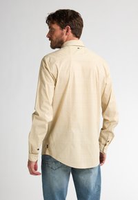 Camisa amarilla clara con un patrón sutil, puños abotonados y dobladillo curvado. Llevada con jeans de mezclilla azul. Sin logotipos prominentes visibles.