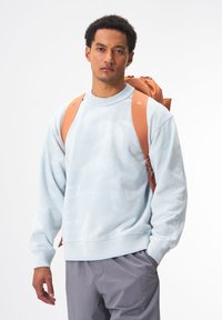 Hellblaues Tie-Dye-Sweatshirt mit gerippten Bündchen und Saum, kombiniert mit einem braunen Rucksack mit gepolsterten Schultergurten und einem Kordelzugverschluss.