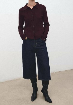 Personne portant un cardigan boutonné de couleur bordeaux foncé, un pantalon culotte en denim foncé à jambes larges, et des bottes noires à talons pointus, debout les mains dans les poches.