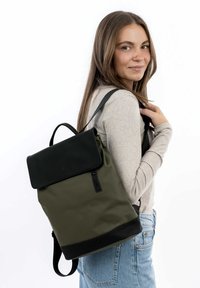 Grüner Stoff-Rucksack mit einer Klappe aus schwarzem Leder, rechteckiger Form, verstellbaren Trägern und einem oberen Griff. Glatte Textur mit minimalistischem Design.