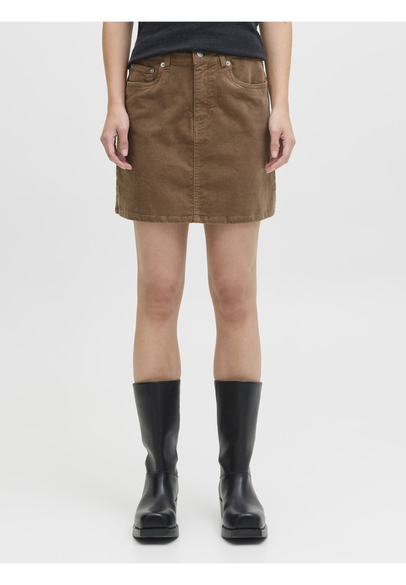 Femme portant une jupe en velours côtelé marron, un haut noir et des bottes noires mi-mollet, debout devant un fond uni.