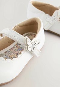 Zapatos de charol blancos para niños con cierres de tira en forma de mariposa y plantillas impresas con flores sobre un fondo claro.