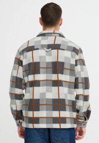 Camisa chaqueta de forro polar a cuadros grises y blancos con acentos naranjas, un cuello con solapa y un lazo en la parte posterior para colgar.