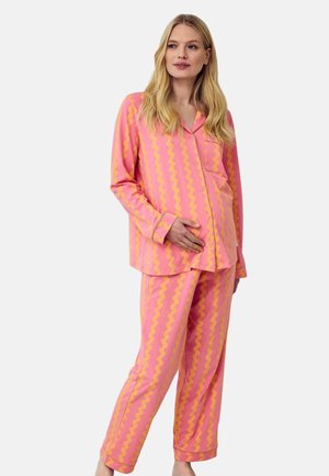 Gravid kvinde iført lyserødt og gult, bølget, stribet pyjamasæt med lange ærmer, der forsigtigt holder sin mave, stående mod en hvid baggrund.