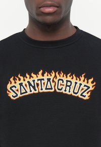 Zwart sweatshirt met "SANTA CRUZ" in vet, witte en oranje vlam-geborduurde letters. Klassiek ontwerp met ronde hals.