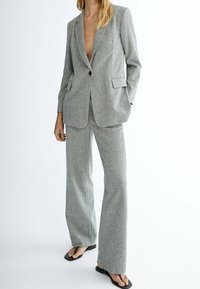 Kobieta w jasnoszarej dopasowanej marynarce i pasujących szerokich spodniach, połączonych z czarnymi sandałami na odkrytej pięcie, stoi z jedną ręką w kieszeni.