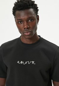 Sorte bomulds t-shirt med hvid broderet "amour" tekst på fronten, rund halsudskæring og korte ærmer. Glat tekstur.