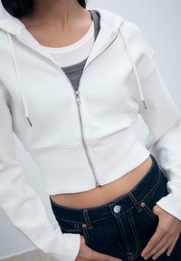 Biely skrátený hoodie s zipsom a šnúrkami, vrstvený cez šedý dámsky nátelník. Nosí sa s tmavými denimovými džínmi so zapínaním na gombík.