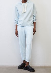 Felpa azzurra chiara con tasca frontale e cappuccio con coulisse, abbinata a pantaloni della tuta coordinati. Scarpe nere chunky senza lacci completano l'outfit.