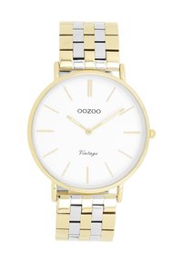 Montre en métal doré et argenté avec un cadran rond blanc, des index horaires minimalistes, le nom de la marque "OOZOO" et des détails en écriture vintage.