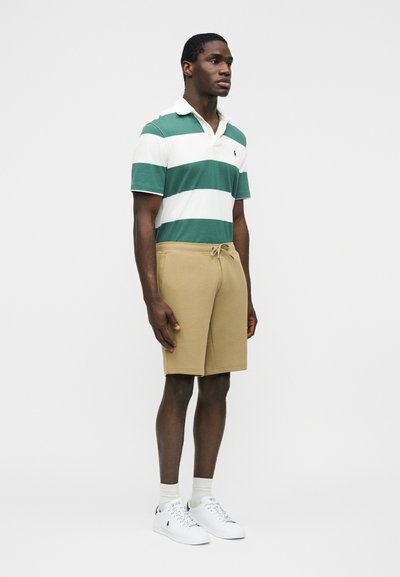 Άνδρας που στέκεται, φοράει πράσινο και λευκό ριγωτό πόλο πουκάμισο, μπεζ σορτς με κορδόνι, λευκές κάλτσες και λευκά αθλητικά παπούτσια μπροστά σε απλό φόντο.