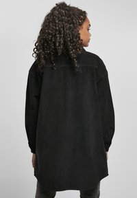 Femme aux cheveux bouclés tournée de profil, portant une longue veste noire en velours côtelé sur un fond clair uni.