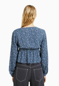 Blusa floreale blu con maniche lunghe, vita arricciata e scollatura rotonda. Tessuto texturizzato che presenta delicati motivi di fiori bianchi.