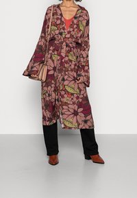 Bloemenpatroon, lange mouwen badjas in roze, groen en burgundy met een strik in de taille. Gecombineerd met een rode top en zwarte broek, met bruine enkellaarsjes.