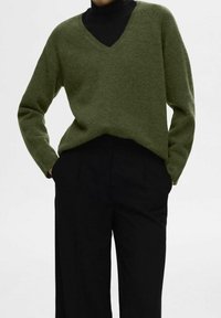 Maglione verde a scollo a V realizzato in materiale morbido, con maniche lunghe e vestibilità leggermente ampia, abbinato a pantaloni neri su misura.