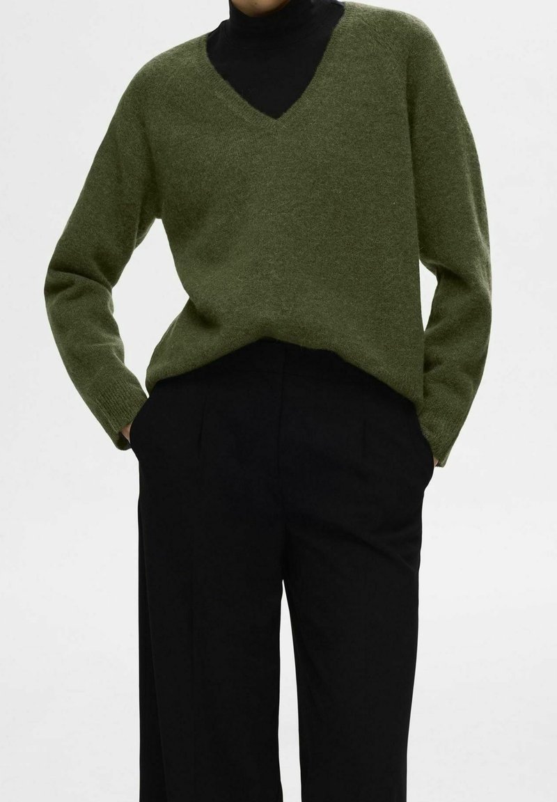 Maglione verde a scollo a V realizzato in materiale morbido, con maniche lunghe e vestibilità leggermente ampia, abbinato a pantaloni neri su misura.