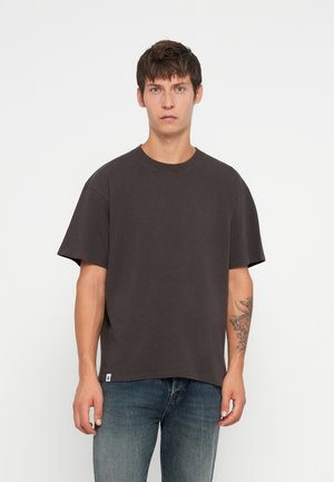 Dunkelgraues T-Shirt mit kurzen Ärmeln, Rundhalsausschnitt und lockerer Passform. Das Material wirkt weich und hat sichtbare Nähte. Ein kleines Logo-Patch am Saum.