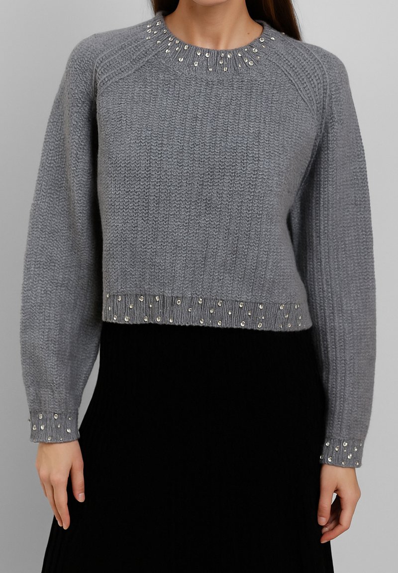 Maglione grigio a maglia con borchie argentate su colletto, polsini e orlo, indossato con una gonna nera, mostrato dalla gola ai fianchi.