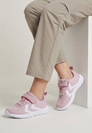 Person iført beige bukser og pinke net-sneakers med hvide chevron-detaljer, siddende på en hvid blok.