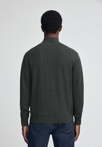 Mörkgrå stickad tröja med hög krage och långa ärmar, har en strukturerad ytfinish och ribbade muddar. Bärs med blå denimjeans.