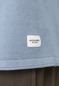Lichtblauwe gebreide trui met een wit label met "JACK & JONES EST. 1990" in zwarte tekst aan de onderkant. Soepele textuur, geribbelde zoom.