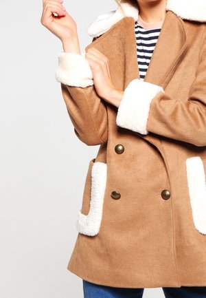 Manteau en laine marron à double boutonnage, col en fausse fourrure blanche avec poches, et boutons en laiton, porté par-dessus un haut rayé.