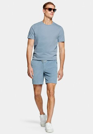 Homme portant un t-shirt bleu clair, un short assorti, des baskets blanches et des lunettes de soleil, marchant devant un fond blanc uni.