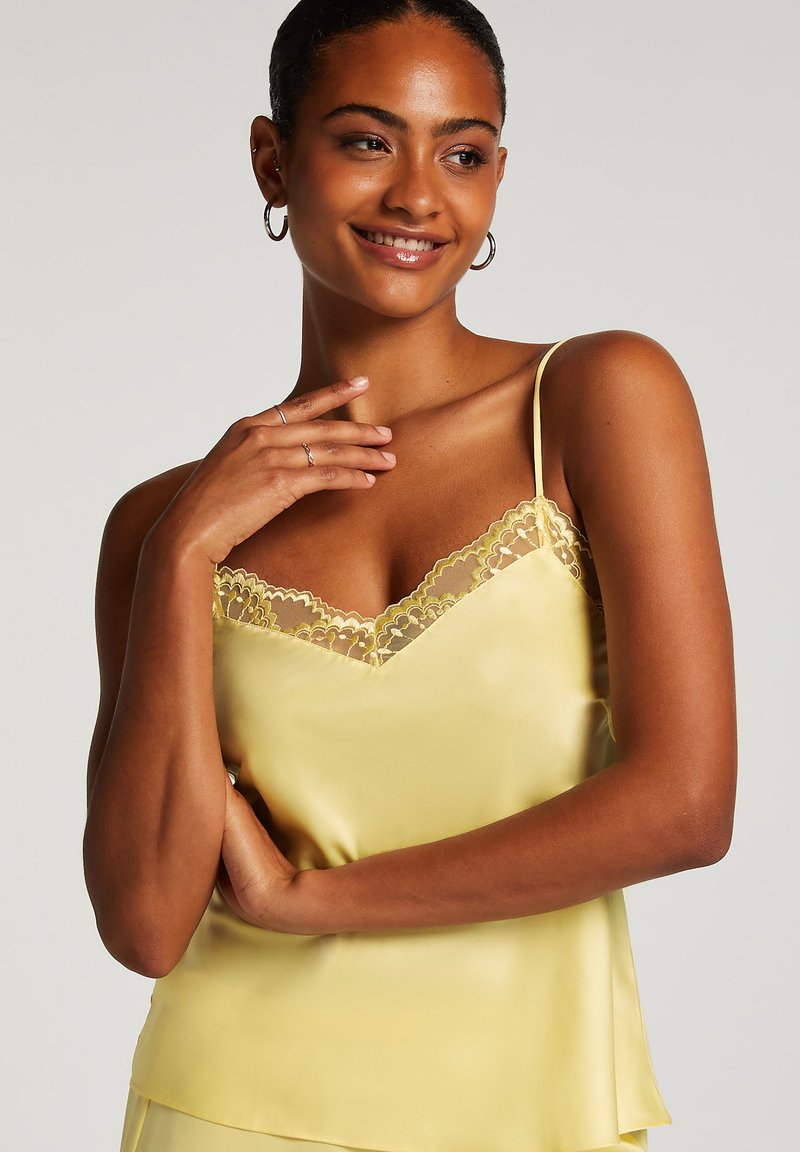 Gult satin camisole med spetskant längs urringningen. Har justerbara band och en mjuk textur. Visas med en modell.