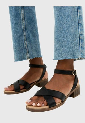 Piedi che indossano sandali neri in pelle con cinturini alla caviglia e tacco basso in legno, abbinati a jeans corti di denim blu.