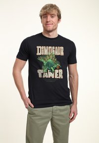 Hombre sonriente con cabello rubio despeinado, vistiendo una camiseta negra con un estegosaurio verde y el texto "Domador de dinosaurios", y pantalones caqui.