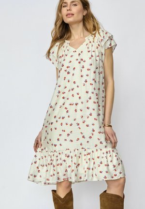 Žena oblečená v bielych šatách po kolená s červeným a fialovým kvetinovým vzorom a volánikmi na leme, doplnená hnedými semišovými čižmami strednej výšky po lýtka.