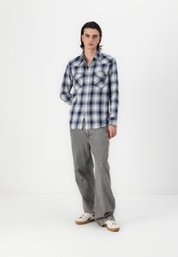 Camicia di flanella a scacchi blu e bianca con due tasche sul petto, abbinata a pantaloni grigi a righe e sneaker bianche su uno sfondo semplice.