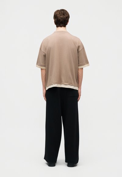 Άτομο που στέκεται με την πλάτη γυρισμένη, ντυμένο με ένα χαλαρό μπεζ πουκάμισο με κοντά μανίκια πάνω από ένα πιο ανοιχτό πουκάμισο και φαρδιά μαύρη παντελόνια σε απλό φόντο.