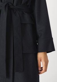 Navy pinstripe coat with a tie belt, wide sleeves, and a patch pocket. Made of a smooth, textured fabric with a tailored design.