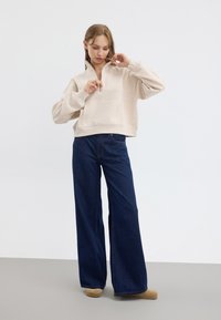 Haut de survêtement beige court avec un demi-zip, poche avant et manches larges, assorti à un jean large bleu foncé et des chaussures à enfiler beiges.