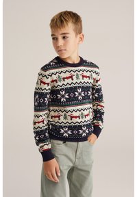 Stickad tröja med krämfärgad bakgrund och marinblå detaljer; har jöliga mönster med röda rävar, gröna träd och vita snöflingor.