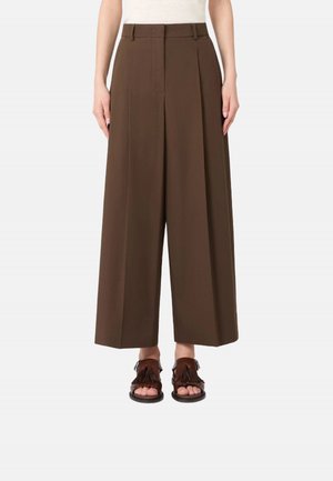 Pantaloni marroni a gamba larga e taglio corto indossati con sandali marroni con nappine e punta aperta su una persona in piedi davanti a uno sfondo semplice.